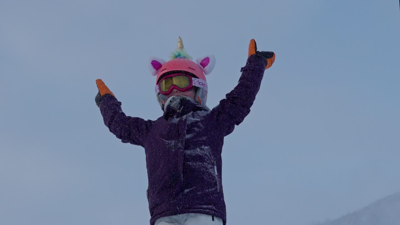 niña de 7 años con casco de unicornio levanta las manos, aplaudiendo en la cima de la colina mientras nieva, cámara lenta