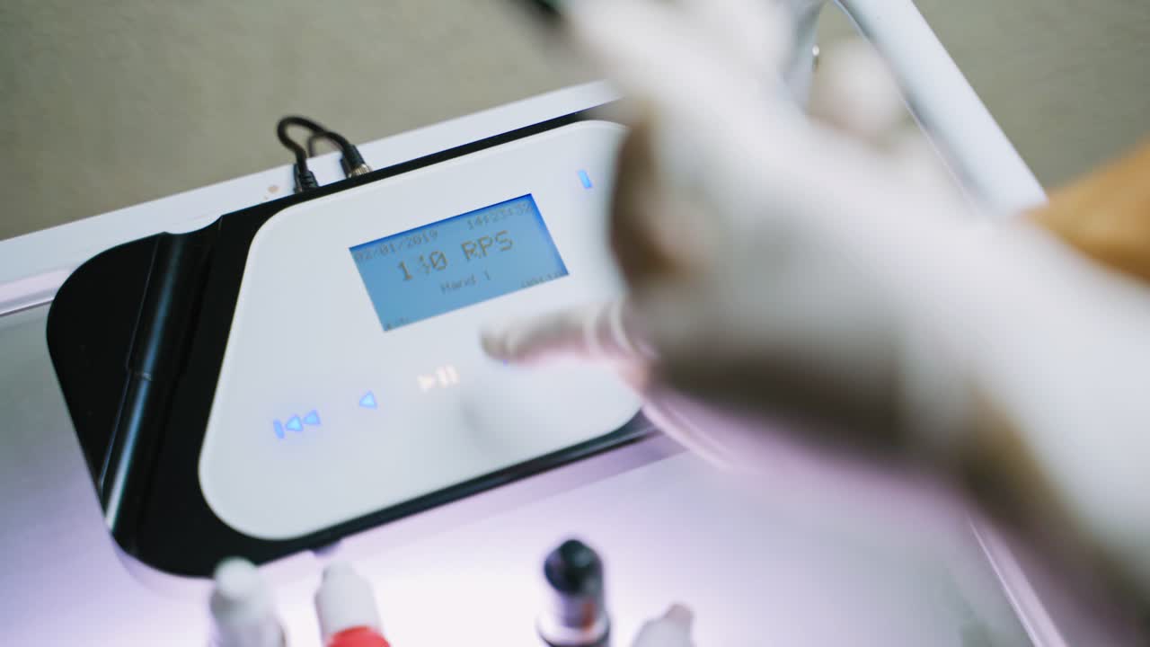 A master in white gloves holds the pen in her one hand, and other sets up the electrical apparatus for the permanent makeup before the procedure. Blurred background.
