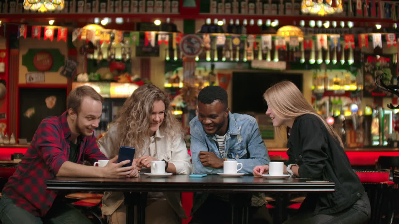 Afro-American man with friends in a cafe laughs and spends time in a cheerful company with a girl watching photos and smiling while telling stories.