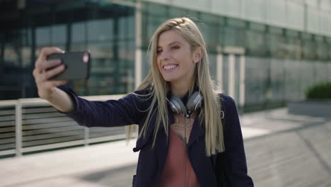 retrato de una joven mujer de negocios rubia y animada, una pasante sonriendo, posando, haciendo caras, tomando una foto selfie usando la cámara de un teléfono inteligente en la ciudad