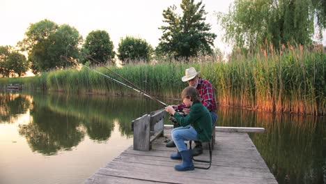 side view of a teen boy sitting with his grandfather on the lake pier, talking and fishing together