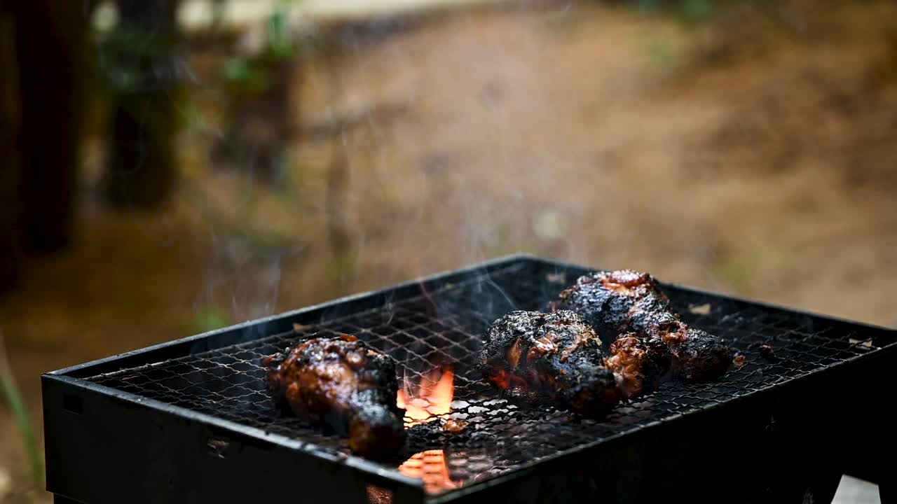 Slow-motion shot of burnt chicken drumsticks cooking over a low flame on a BBQ grill. The sizzling charred meat highlights rustic outdoor barbecue, careful roasting, and reviving overcooked food