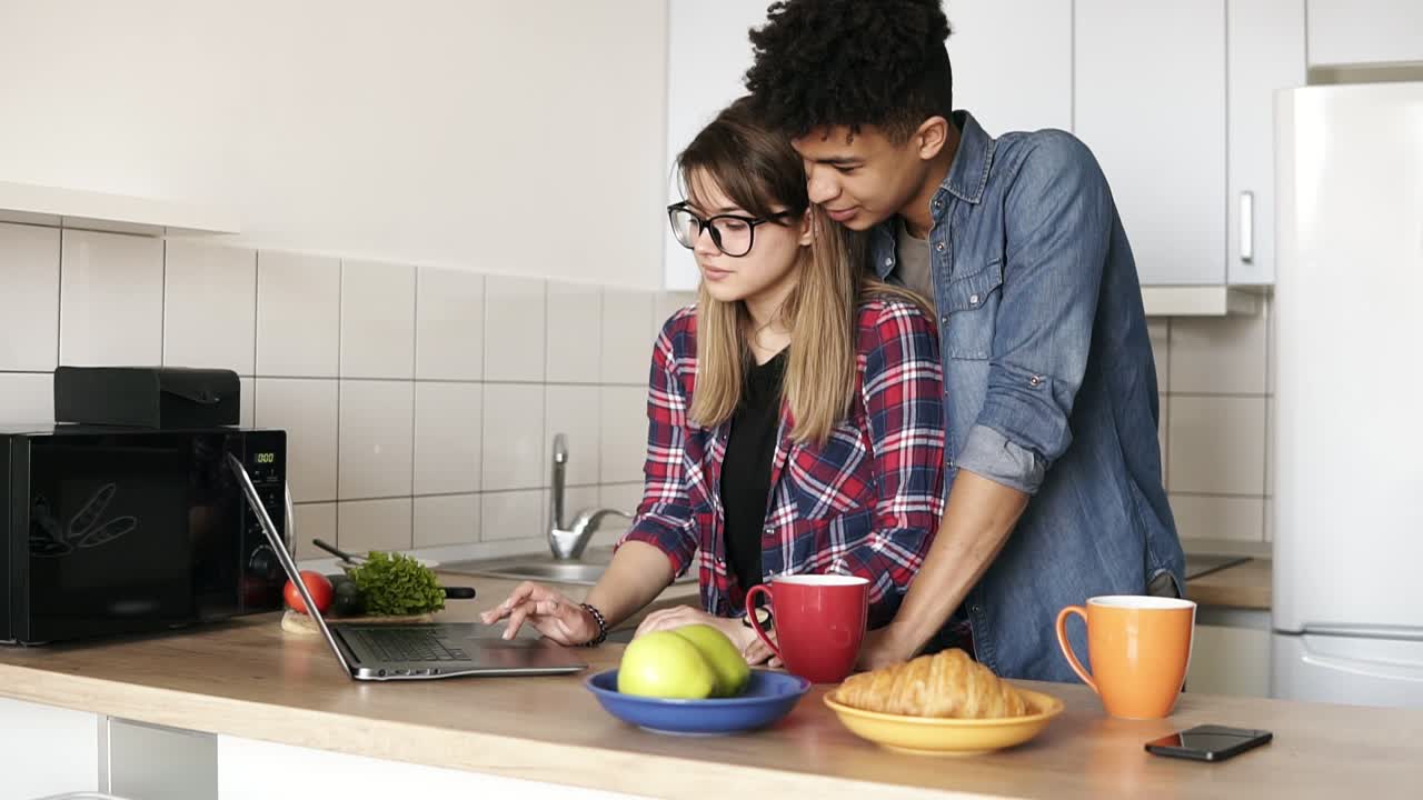 imágenes en cámara lenta de una linda pareja navegando por la web, usando una computadora portátil mientras están sentados en una cocina