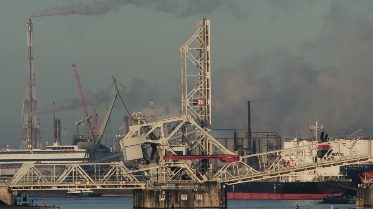 Static shot of a moving bridge, thick smoke rising from chimneys and boat activity in a busy port