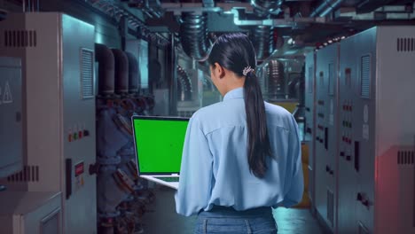 Back View Of An Asian Female Professional Worker Standing In Engine Control Room, Typing On Laptop'S Keyboard With Green Screen And Look At The Factory Once
