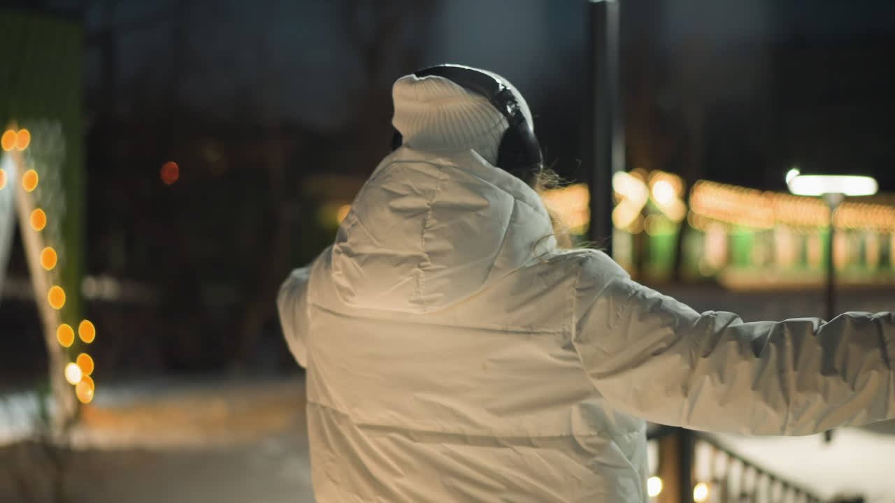 Individual spinning joyfully on snow covered park walkway under warm evening lights wearing white coat beanie and headphones showing expressive movement in winter night scene in ambient urban setting