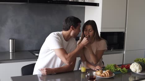 Young-man-and-woman-eating-croissants,-driking-tea-and-talking-sitting-at-table-counter-in-kitchen-together-enjoying-peaceful-morning-at-home-and-feeding-each-other.-Food,-apartment-and-people-concept