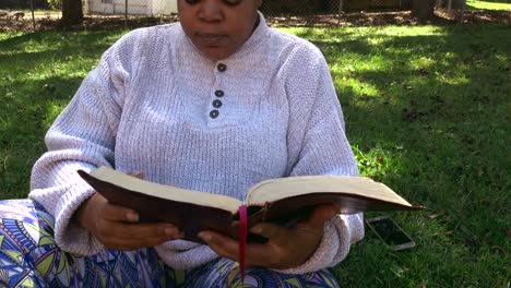 primer plano medio de una mujer negra sentada leyendo la biblia y sin prestar atención a la distracción de su celular en el fondo