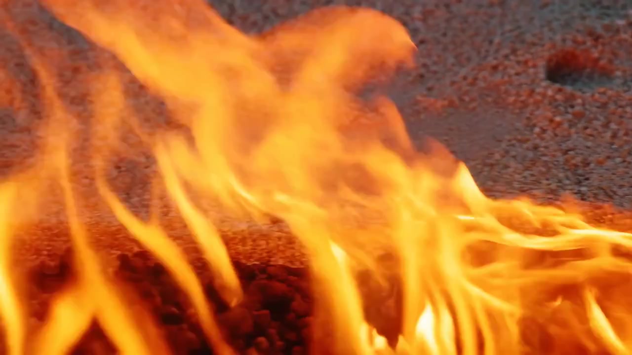 Vista baixa próxima ao chão mostra línguas de chama amarelo-alaranjadas brilhantes subindo de brasas incandescentes e carvões escuros sobre uma superfície de seixos grossos, com desfoque suave de fundo e intenso contraste quente.