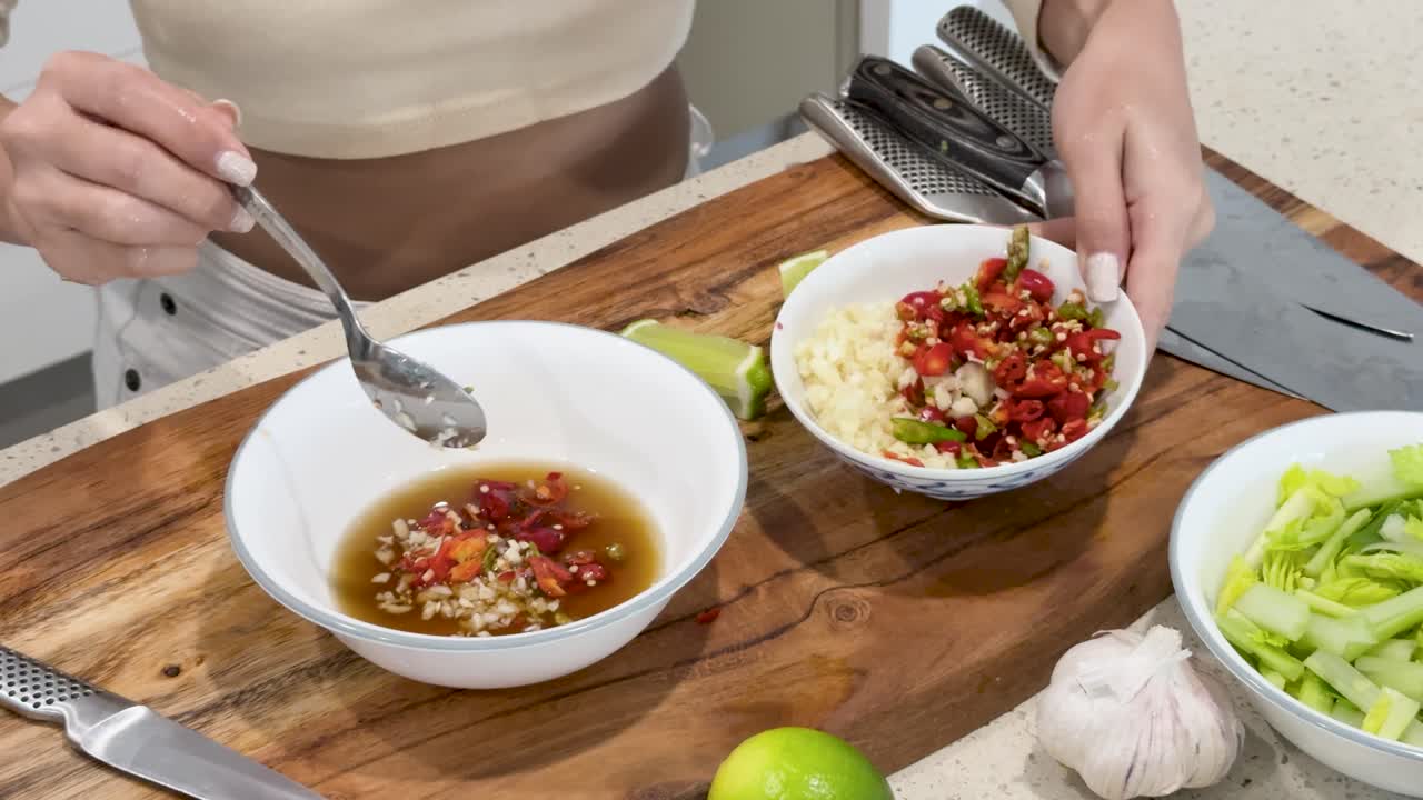 A person prepares a spicy Thai dipping sauce by mixing chopped chili, garlic, and citrus juice in a bowl on a wooden kitchen counter under bright lighting