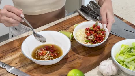 a person prepares a spicy thai dipping sauce by mixing chopped chili, garlic, and citrus juice in a bowl on a wooden kitchen counter under bright lighting