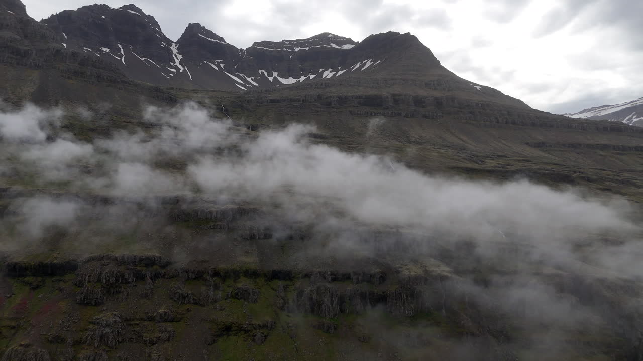 Misty mountain slopes and small waterfalls overlook Berufjörður as drifting clouds slide across the rugged Icelandic landscape