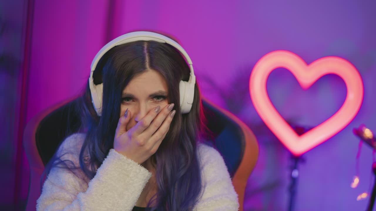 Unhappy young woman wearing white headphones seated in colorful studio with glowing heart light in background, showing visible frustration and disappointment after recording session went wrong