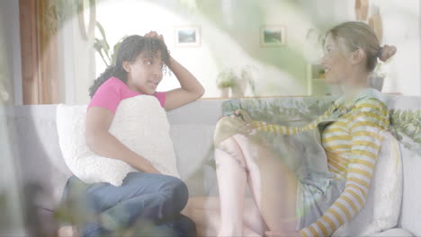 animation of sunlight and plants over two happy diverse teenage girls talking on couch