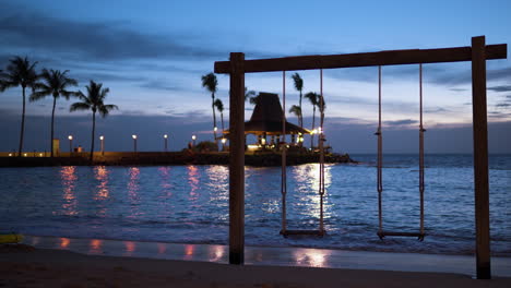 wooden rope swing on tropical beach at shangri-la resort on colorful sundown, sunset bar pavillion in background lit with torches and sunlight