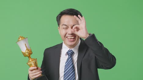 close up of asian business man in a suit and tie with a gold medal and trophy showing okay hand sign over eye and smiling to camera as the first winner on green screen background in the studio