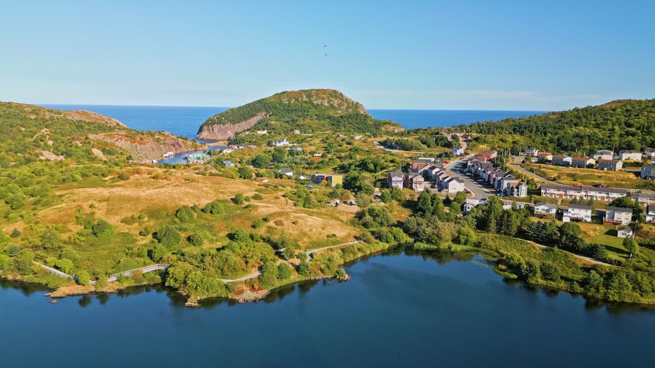 A drone soars over a grassy peninsula at Quidi Vidi, revealing rugged hills and a sheltered harbour with clear blue water and panoramic views of the coastline