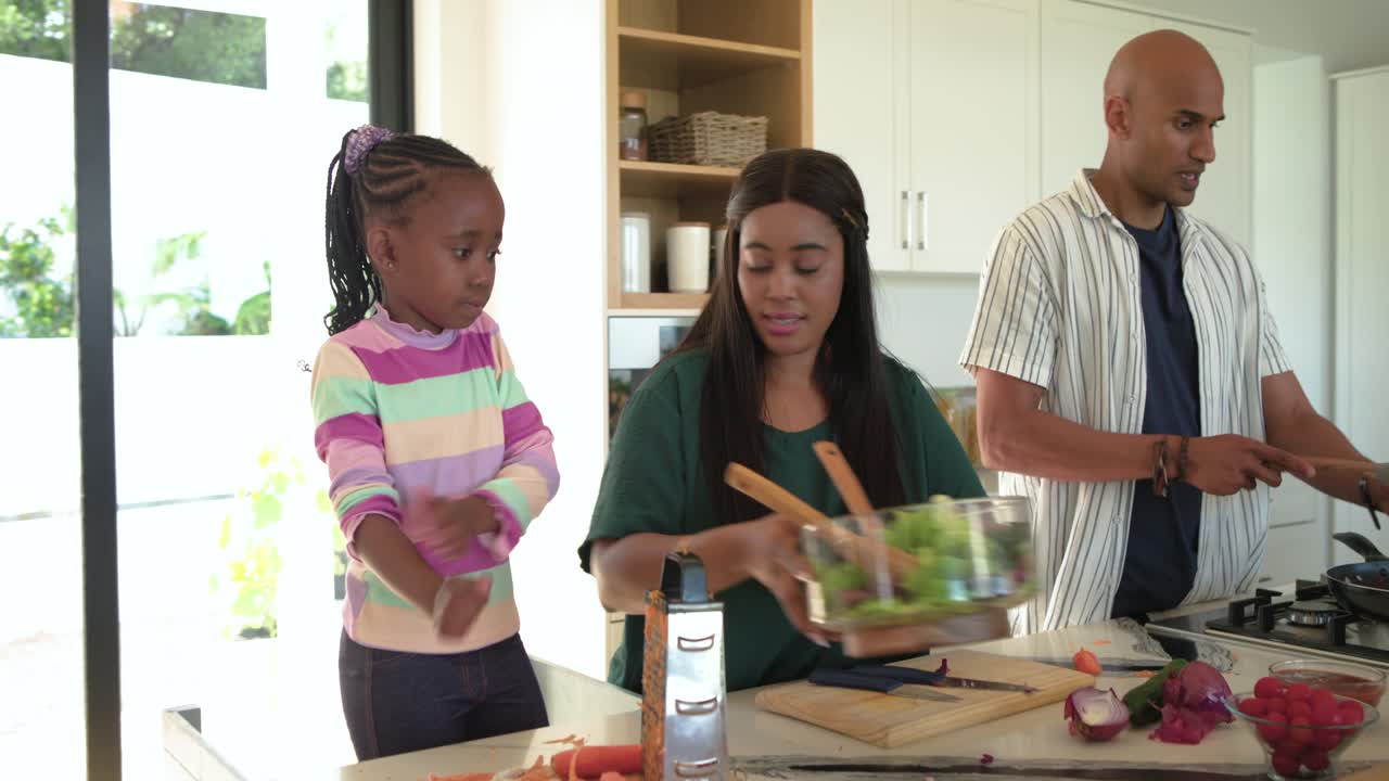 Black mother guiding daughter grating carrots while father flipping veggies on kitchen island