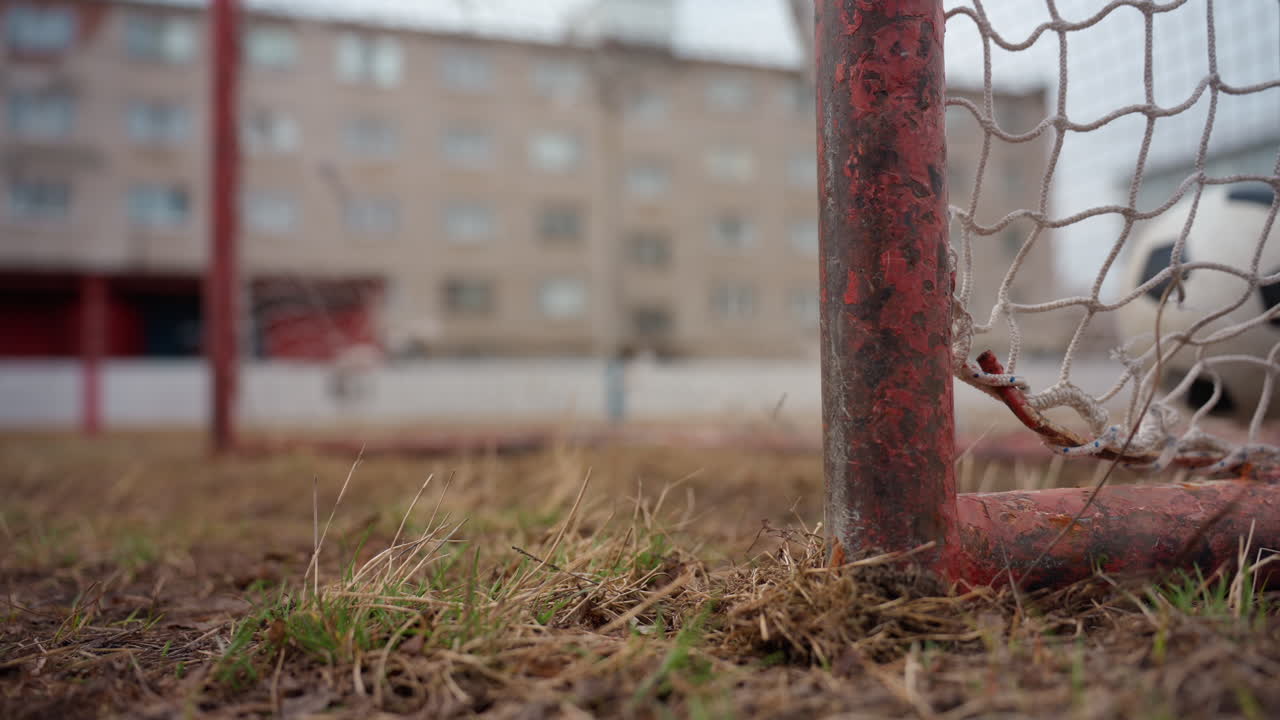 porta da calcio a livello del suolo con erba in primo piano. La prospettiva dal basso mostra un palo arrugginito, una rete aggrovigliata e un terreno fangoso, un pallone vagante che rotola nell'inquadratura, caseggiati di periferia sullo sfondo, promettente base