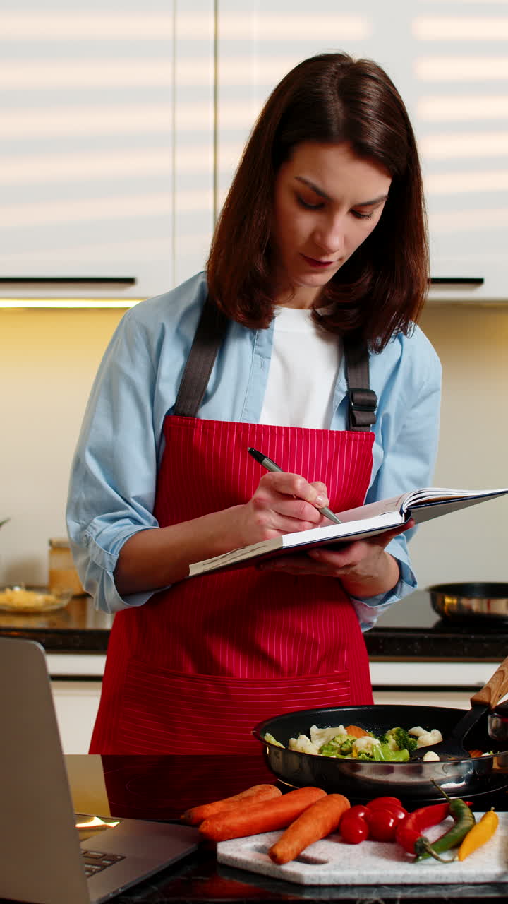 Giovane donna che impara a cucinare sano dal laptop, prendendo appunti, nella moderna cucina di casa.