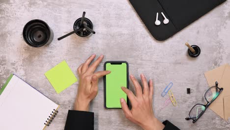 female tapping on smartphone device with green screen, top view of office work desk. woman using smartphone displaying chromakey. female fingers zooming, tapping on smartphone green screen