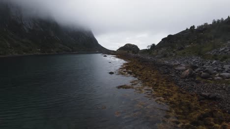 Volando-A-Lo-Largo-De-La-Orilla-Cerca-Del-Agua-Mirando,-A-La-Izquierda-Las-Montañas-Están-Cubiertas-De-Niebla,-En-Cámara-Lenta