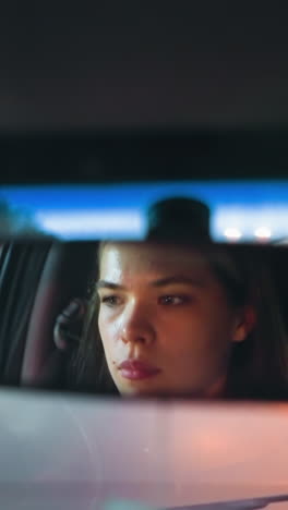 la mujer se refleja en el espejo retrovisor mientras conduce su coche, con la carretera y un coche rojo delante de ella visible en el fondo, sus luces encendidas, mientras se mueve a través de una noche débilmente iluminada