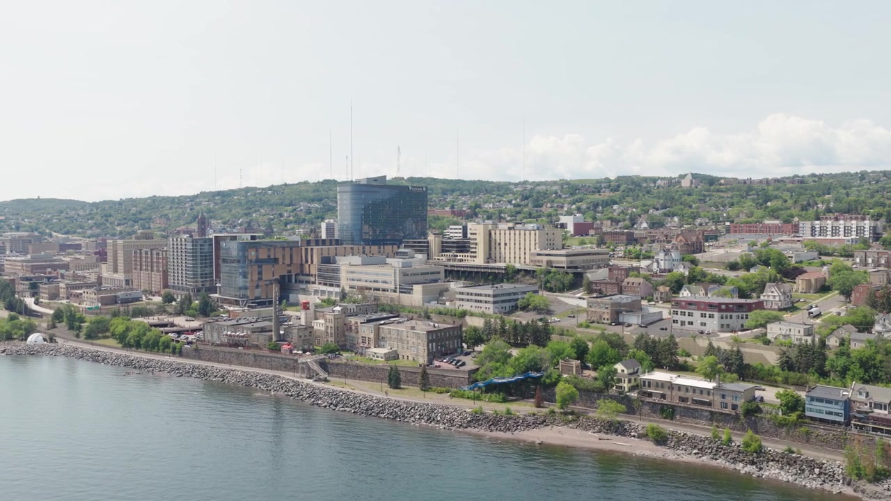 Un drone orbita sullo skyline del centro di Duluth, mostrando moderni edifici medici con torri di vetro e quartieri collinari sopra la costa del Lago Superiore.