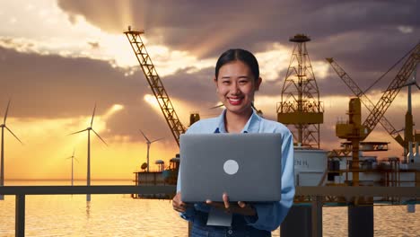 mujer de negocios trabajando en una computadora portátil al atardecer con vistas a la granja eólica y la plataforma petrolera
