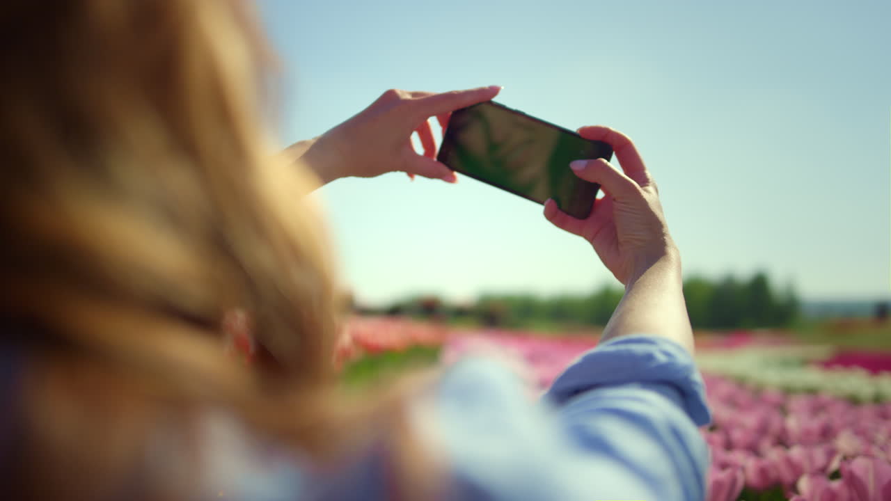 Vista de costas de garota sorridente tirando uma selfie com o celular em um campo de flores.