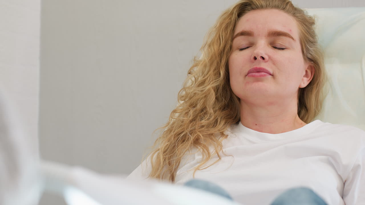 Client in white top sits calmly on clinic chair with eyes closed in relaxed posture, appearing to gently open eyes under soft lighting, conveying peace and personal care atmosphere
