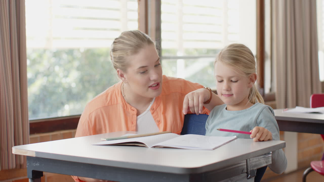 In school, female teacher smiling and helping young student with homework at desk