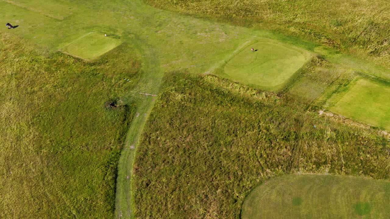 Drone captures golfers walking fairway on sunny day, revealing lush green landscape and bunkers