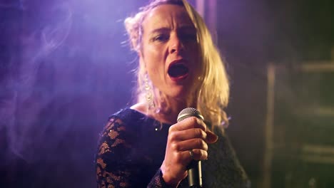 handheld medium shot of a talented blonde singer performing and singing a song emotionally during a concert with a coloured light show in changing colours