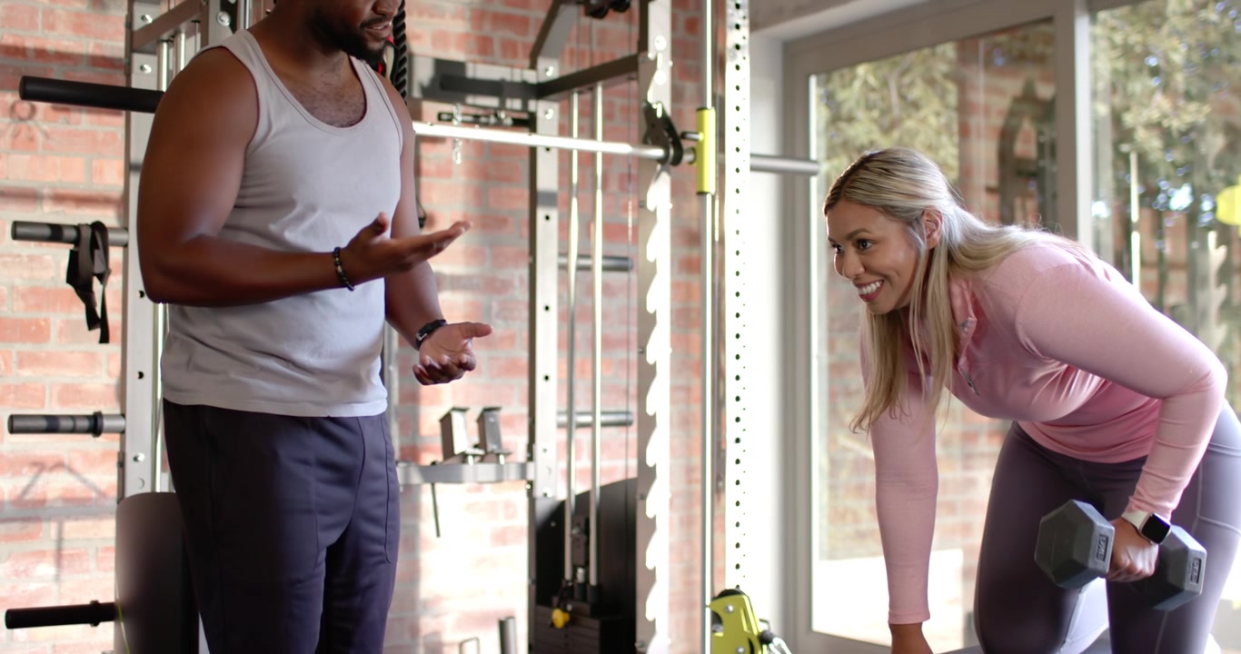 African American trainer giving cues, woman client pulling hex dumbbell on bench, correcting form