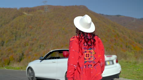 woman in red jacket and white hat by a white convertible car in the mountains