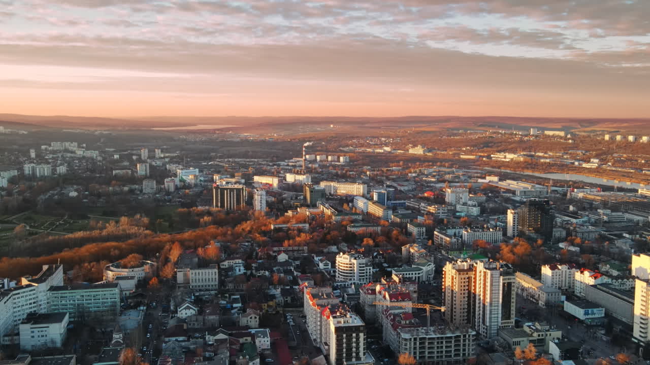 Luftaufnahme von Chisinau bei Sonnenuntergang. Panoramaansicht von mehreren Gebäuden, Straßen mit fahrenden Autos, kahlen Bäumen, Park, See und Hügeln in der Ferne. Moldawien