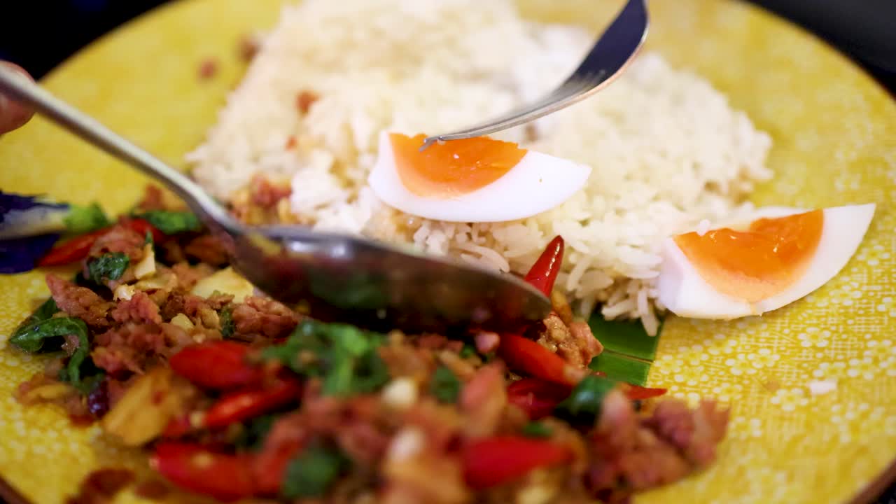 Close-up sequence of a hand using a spoon to mix steamed rice, boiled egg, and fermented pork sausage with herbs and chili on a yellow plate under bright lighting
