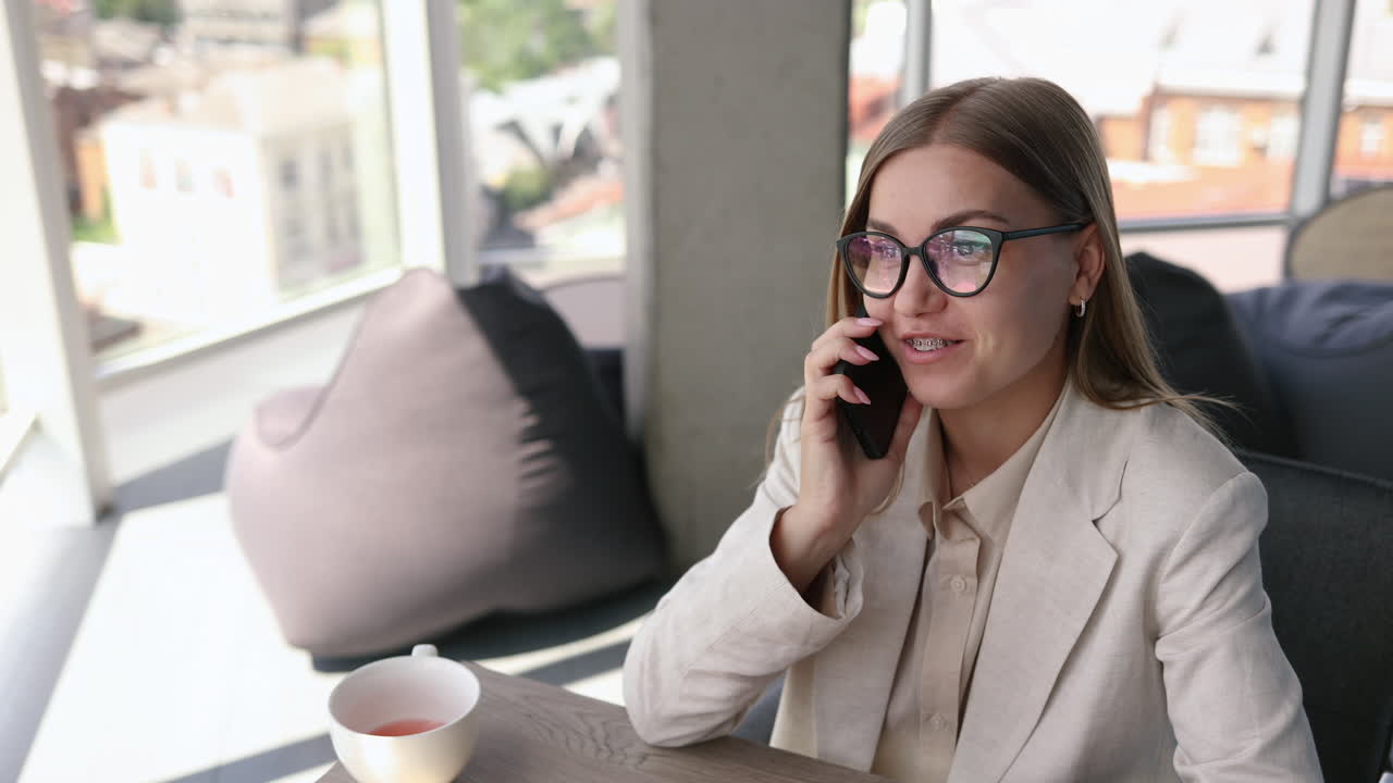 Bella giovane donna con gli occhiali che lavora in un ufficio luminoso e beve tè al laptop. Affascinante signora dai capelli lunghi con gli occhiali che parla al telefono