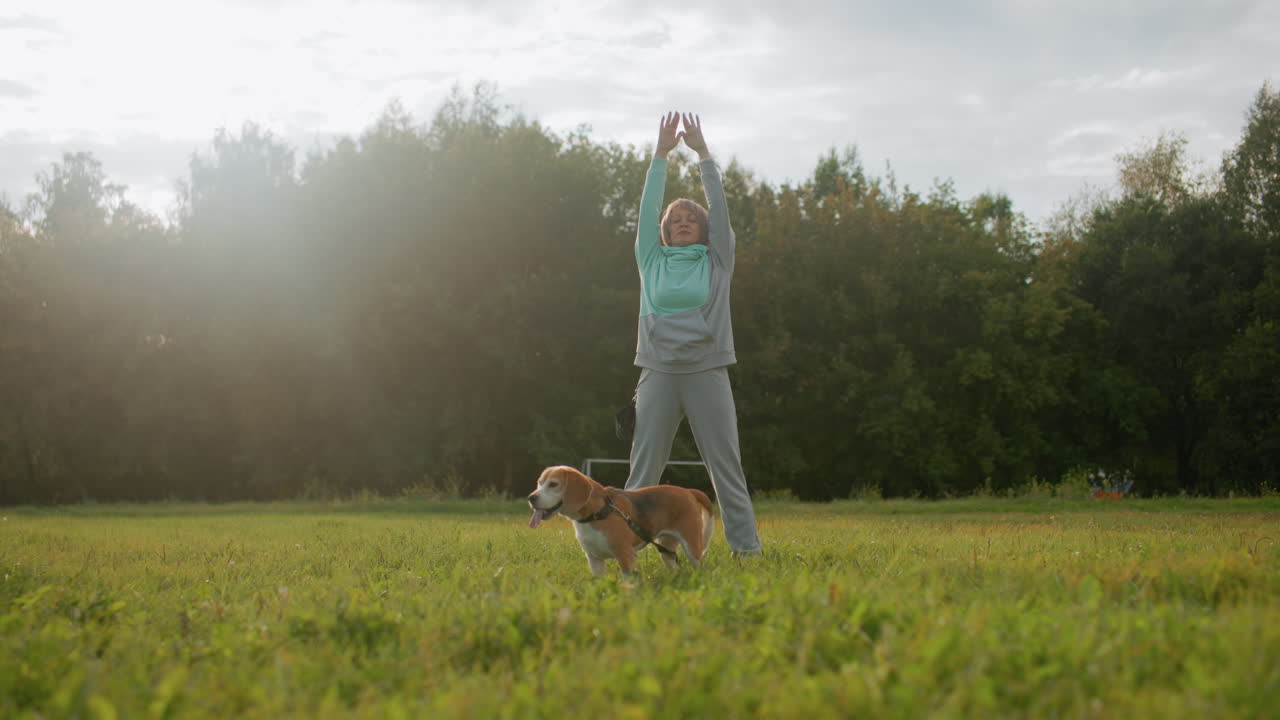 Canine specialist wearing mint tracksuit standing on football field raising hands up and down during outdoor morning workout session under cloudy sky accompanied by beagle dog