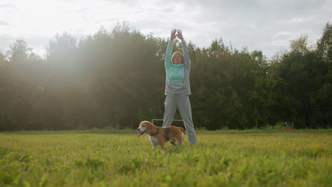 canine specialist wearing mint tracksuit standing on football field raising hands up and down during outdoor morning workout session under cloudy sky accompanied by beagle dog