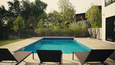 push in shot over black pool chairs to a large blue pool in the backyard of a luxury home