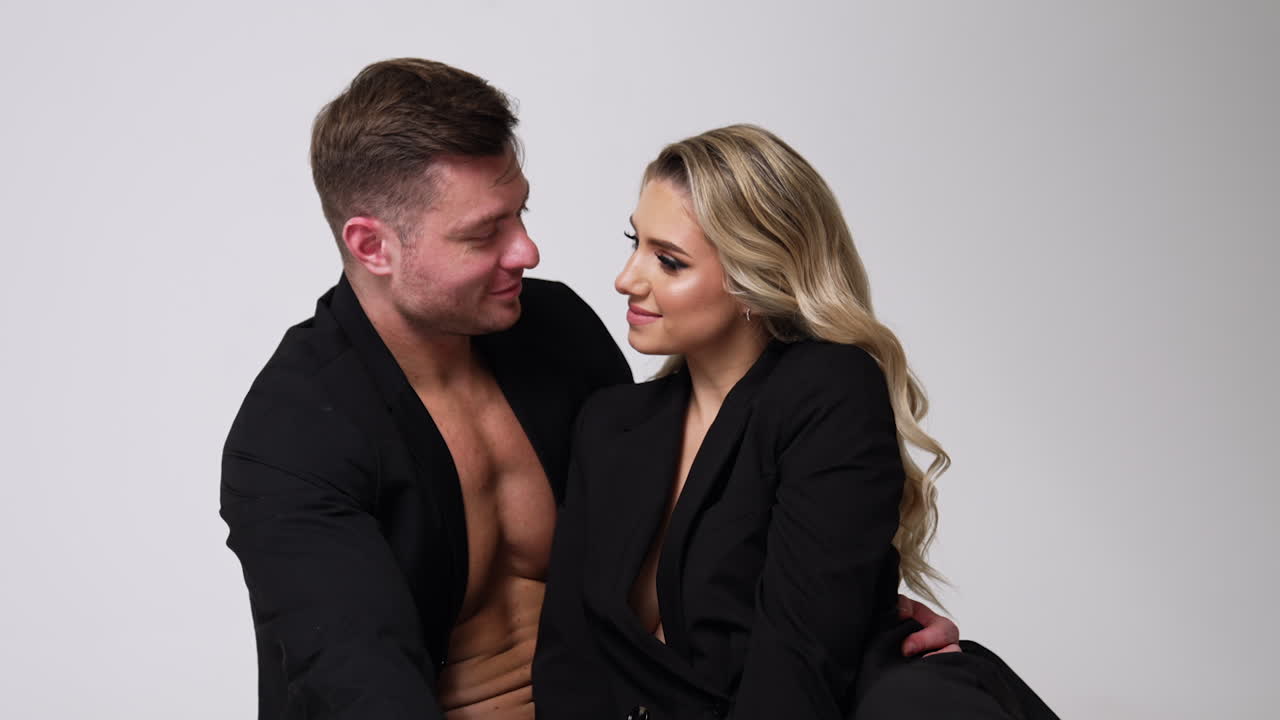 Beautiful Caucasian couple both wearing the black jackets. Man and woman sit close together looking at each other. White backdrop.