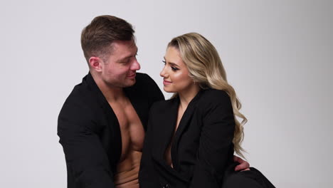 beautiful caucasian couple both wearing the black jackets. man and woman sit close together looking at each other. white backdrop.