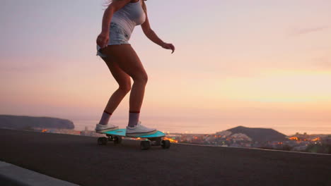 Against-the-backdrop-of-a-sunset,-a-young-and-stylish-skateboarder-rides-her-board-in-shorts-on-a-mountain-road,-showcasing-the-mountains'-enchanting-view-in-slow-motion