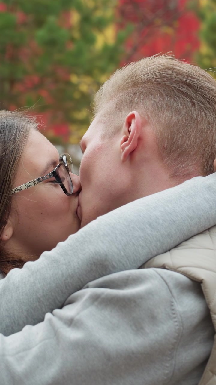 Couple embraces closely in natural setting with greenery. Faces remain focused in tight frame emphasizing intimacy. Blurred green and red foliage creates natural backdrop