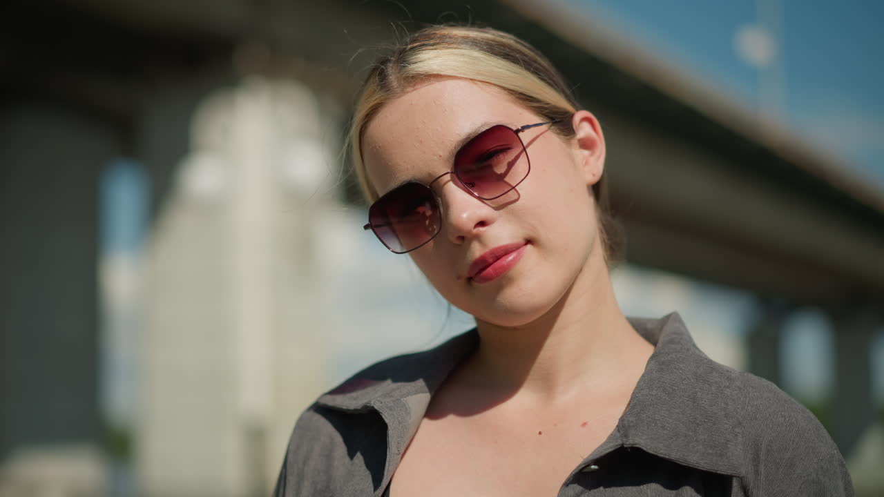 женщина в серой рубашке и солнцезащитных очках наклоняет голову слегка вправо, улыбаясь, она выглядит сосредоточенной, находясь на открытом воздухе, с размытым фоном с мостом и ярким небом