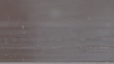 Close-up-shot-of-rain-drops-falling-on-a-wooden-table-making-splashes-with-chairs-in-the-background-in-slowmotion-LOG