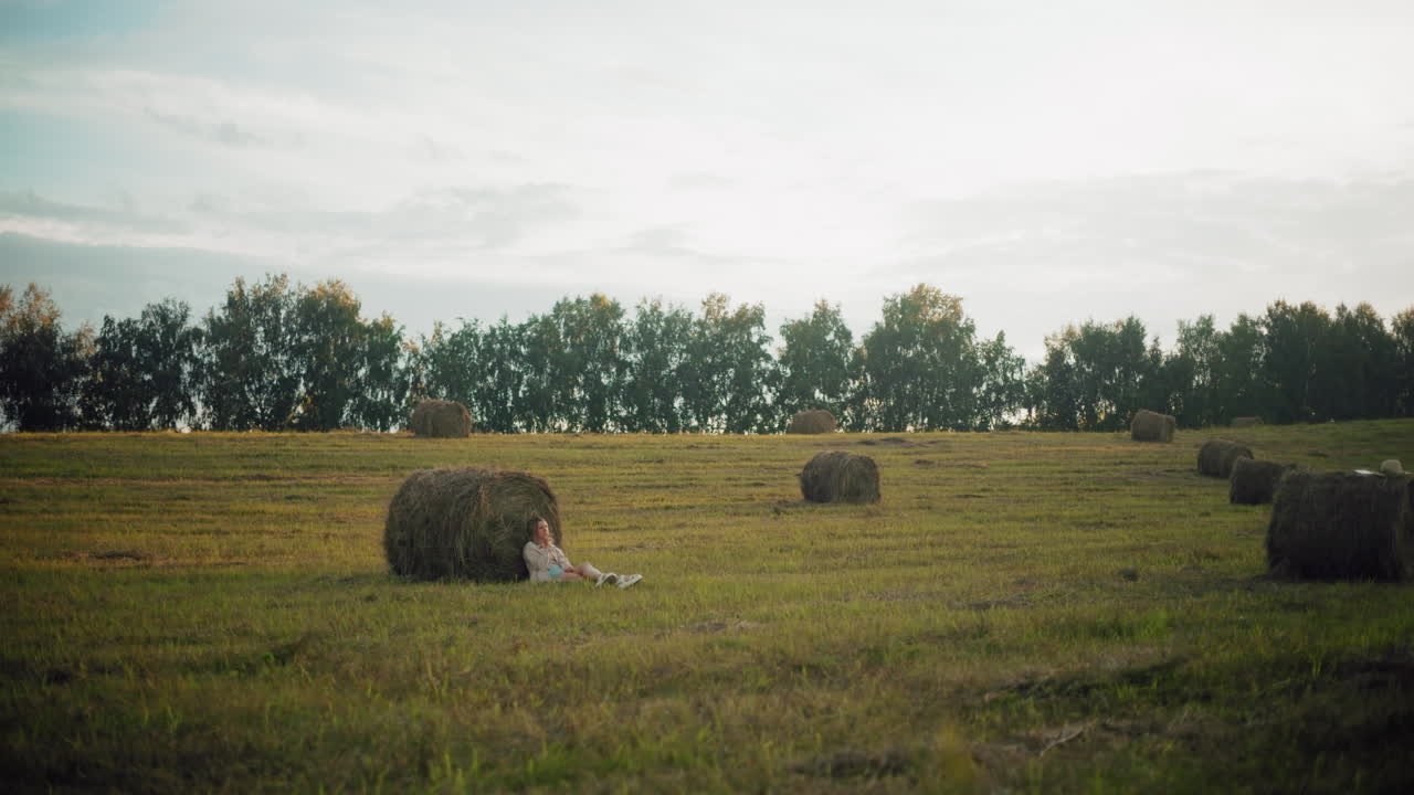 vista distante de la dama sentada en el suelo, descansando contra una gran bala de heno en tierras de cultivo abiertas, paisaje rural pacífico con balas de heno esparcidas y luz cálida de la tarde