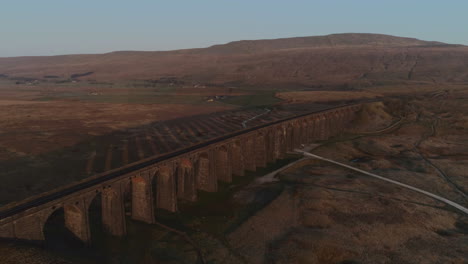 Toma-Aérea-De-Drones-Tirando-Hacia-Atrás-Del-Puente-Del-Tren-Del-Viaducto-Ribblehead-Con-Una-Larga-Sombra-En-El-Impresionante-Amanecer-En-Verano-En-Los-Valles-De-Yorkshire-Inglaterra-Reino-Unido-Con-3-Picos-De-La-Montaña-Whernside-En-El-Fondo